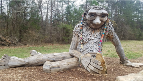 A large wooden sculpture of a seated figure with colorful decorations, surrounded by trees and grass.