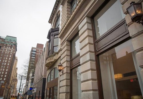 A street view of a historic building with large windows and decorative architecture, alongside modern structures.