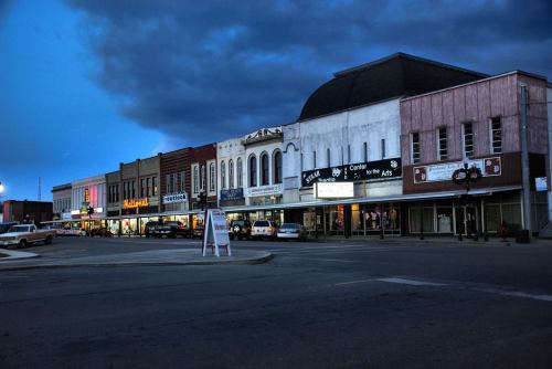 A quiet downtown street at dusk, featuring historic buildings and storefronts under a cloudy sky.