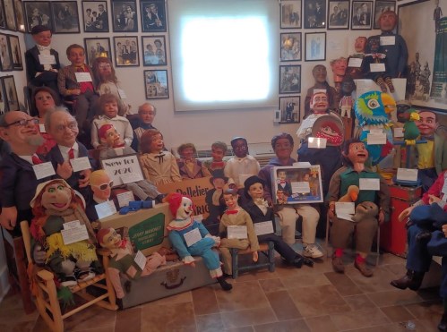 A collection of ventriloquist dummies and puppets displayed in a room filled with framed photos and memorabilia.