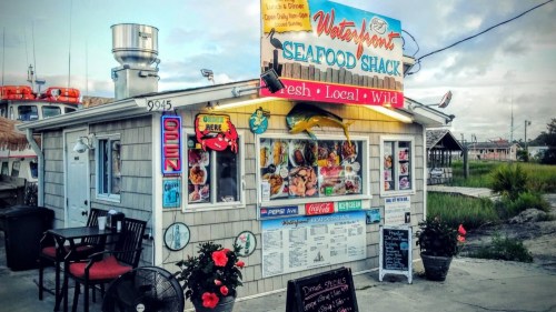 A colorful seafood shack with a sign reading "Waterfront Seafood Shack," featuring outdoor seating and a menu display.