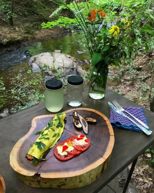 A wooden platter with a colorful omelet, tomatoes, and mushrooms, set on a table by a stream with flowers in a vase.