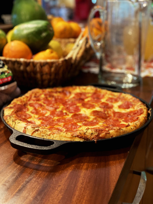 A freshly baked pepperoni pizza in a cast iron skillet, with a fruit basket in the background.
