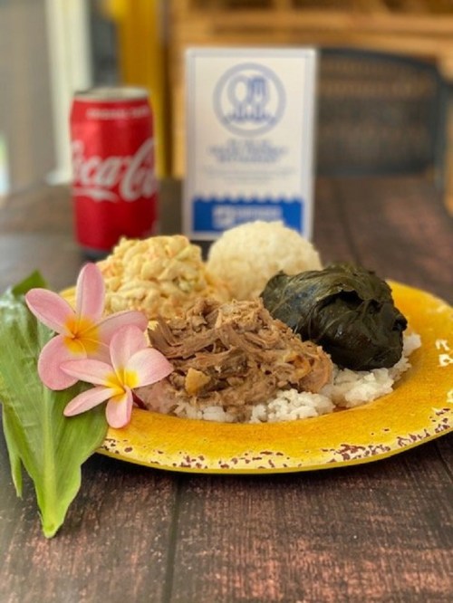 A colorful plate of rice topped with shredded meat, wrapped leaves, and a side of salad, with a soda and flowers nearby.