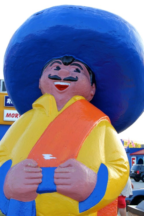 A colorful, oversized statue of a smiling man wearing a large blue sombrero and a bright yellow outfit.