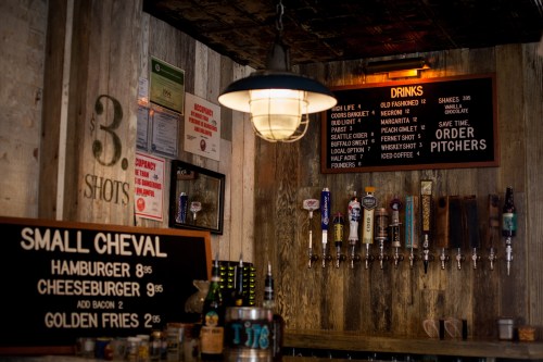 A rustic bar interior featuring a menu board with drinks and food prices, illuminated by a hanging light fixture.