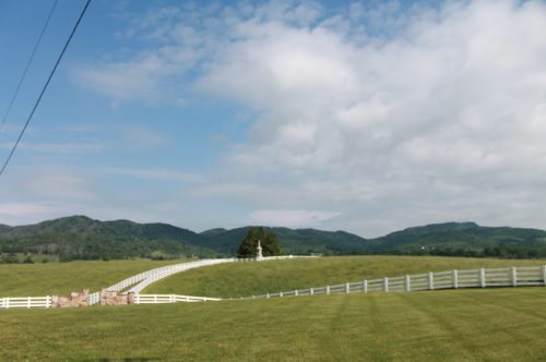 A scenic landscape featuring rolling green hills, a white fence, and a cloudy blue sky.