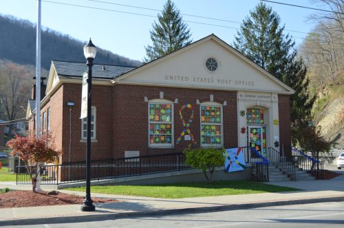 A brick building with colorful window decorations, labeled "United States Post Office," surrounded by trees and a sidewalk.