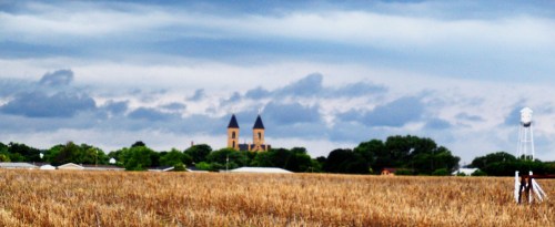 A rural landscape with a golden field, a church with twin spires in the background, and a water tower nearby under cloudy skies.