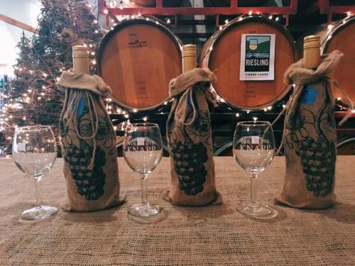 Four wine bottles wrapped in burlap sit on a table with glasses, surrounded by festive lights and wooden barrels.