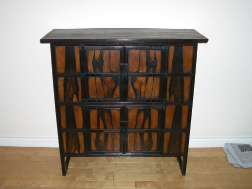 A wooden cabinet with a dark top and patterned brown doors, resting on a light wooden floor against a plain wall.