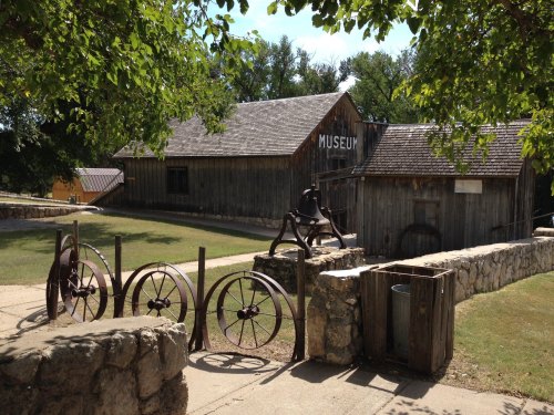 A rustic museum building surrounded by greenery, featuring old wheels and a stone wall in the foreground.