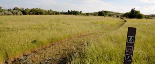A grassy field with a dirt path, marked by signs for walking and biking, under a clear blue sky.