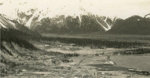 A black-and-white aerial view of a valley surrounded by mountains, with a small town and a body of water visible.