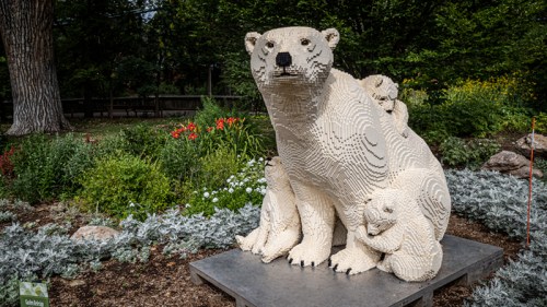 A large polar bear sculpture made of LEGO bricks, surrounded by smaller bear cubs, set in a garden with flowers.