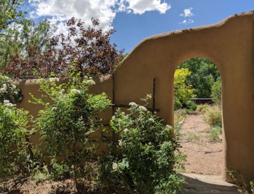 A rustic adobe wall with an archway, surrounded by greenery and colorful plants under a bright blue sky.