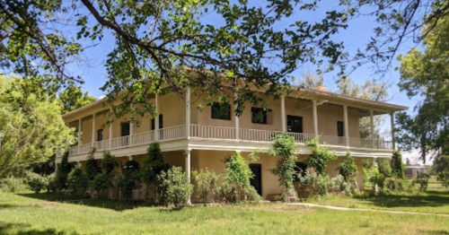 A two-story house surrounded by greenery, featuring a wraparound porch and large windows.