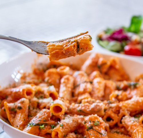 A fork holding a piece of rigatoni pasta with a creamy sauce, next to a bowl of fresh salad.