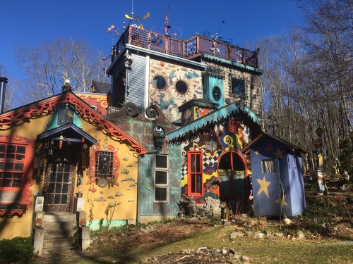 A colorful, whimsical house with unique architectural features and vibrant decorations, surrounded by trees and greenery.