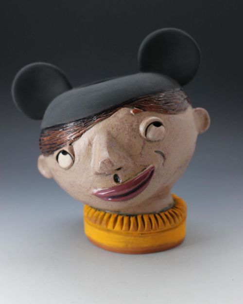 A whimsical ceramic sculpture of a head with mouse ears, a smiling face, and a textured orange collar.