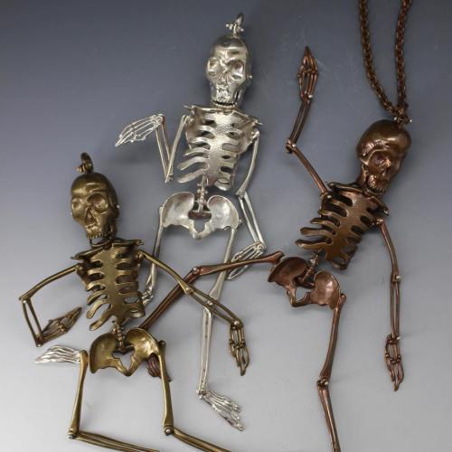 Three decorative skeleton figurines in silver, bronze, and copper, posed playfully against a gradient background.