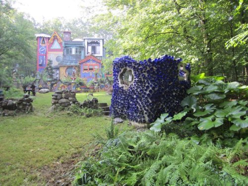 A colorful, whimsical house surrounded by greenery and a unique blue bottle sculpture in the foreground.