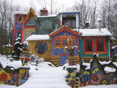 Colorful, whimsical house with unique architecture, surrounded by snow-covered trees and vibrant garden decorations.