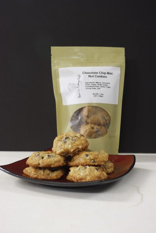 A plate of chocolate chip mac nut cookies next to a packaged cookie bag on a dark background.