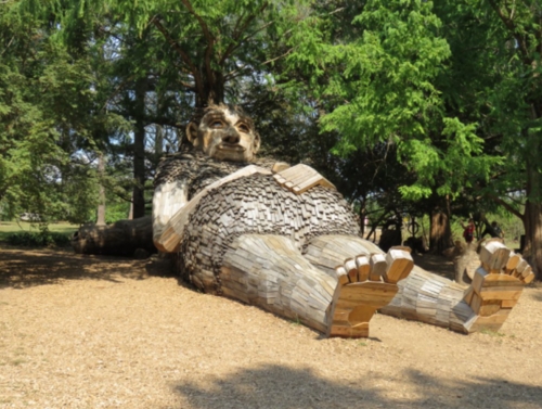 A large wooden sculpture of a reclining giant surrounded by trees in a park setting.