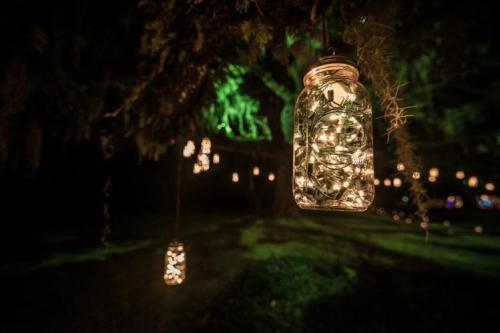 Mason jars filled with fairy lights hang from tree branches, illuminating a dark, green outdoor setting.