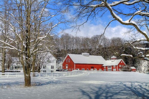 A snowy landscape featuring a red barn, white farmhouse, and bare trees under a blue sky with scattered clouds.