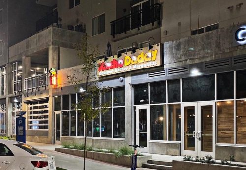 Exterior of a restaurant named "Nacho Daddy" at night, featuring colorful signage and large windows.