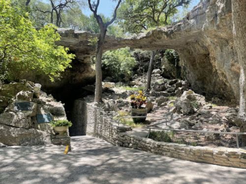 A scenic view of a rocky area with trees, featuring a natural arch and a pathway leading to a cave entrance.