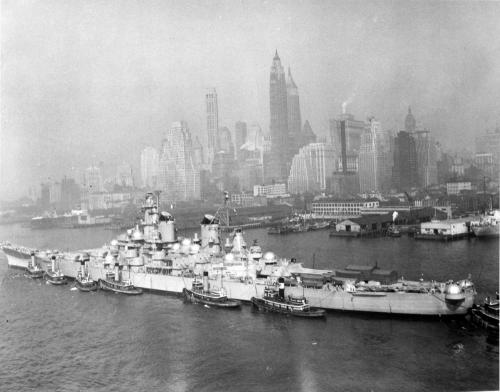 A historic black-and-white photo of a battleship docked near a city skyline shrouded in fog.