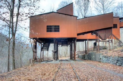 Abandoned industrial building with rust-colored metal exterior, surrounded by trees and fallen leaves on a gravel path.