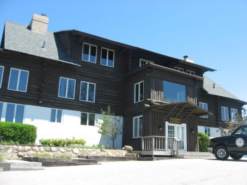 A large log cabin-style building with multiple windows and a front porch, surrounded by greenery and a clear blue sky.