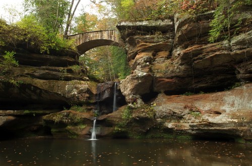 A serene landscape featuring a stone bridge over a rocky area with a waterfall and a calm pool surrounded by trees.