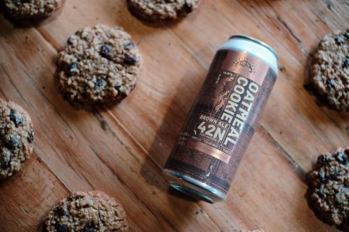 A can of Oatmeal Cookie Brown Ale surrounded by chocolate chip cookies on a wooden surface.