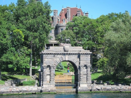 A stone archway leads to a grand mansion surrounded by lush greenery and trees under a clear blue sky.