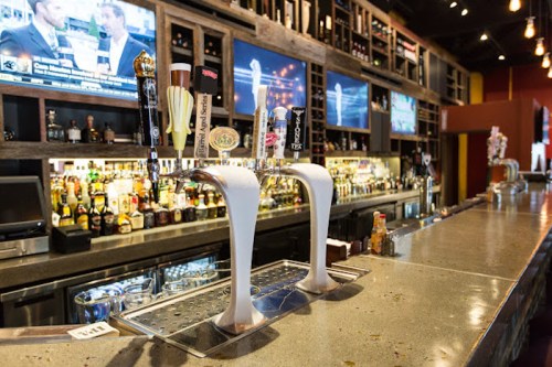 A bar with a variety of beer taps in front of a well-stocked shelf of liquor and multiple screens displaying sports.