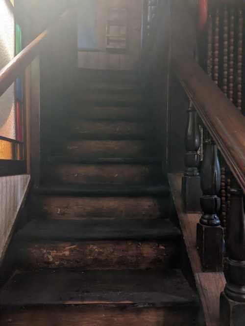 A dimly lit staircase with worn wooden steps and a wooden railing, leading upward in a vintage setting.
