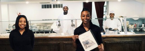 A smiling woman holds a certificate in front of a restaurant counter, with two chefs in white uniforms behind her.