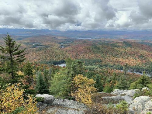 A panoramic view of colorful autumn foliage covering rolling hills and a river, under a cloudy sky.