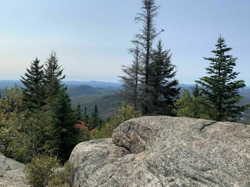 A rocky outcrop overlooks a vast landscape of mountains and trees under a clear sky.
