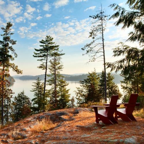 Two red chairs on a rocky outcrop overlooking a serene lake surrounded by trees and a clear blue sky.