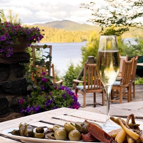 A glass of sparkling wine beside a plate of olives and pickles, with a scenic lake and mountains in the background.