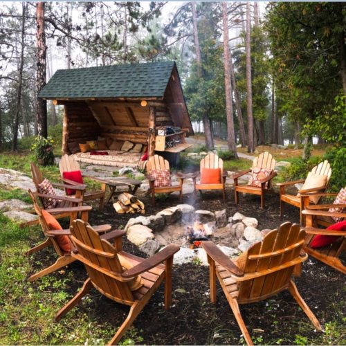 Cozy outdoor seating around a fire pit, with a log cabin and trees in the background. Perfect for gatherings.