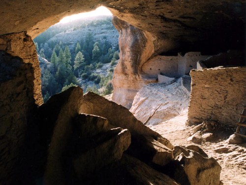 View from a cave entrance, revealing a rocky landscape and ancient stone structures nestled among trees.