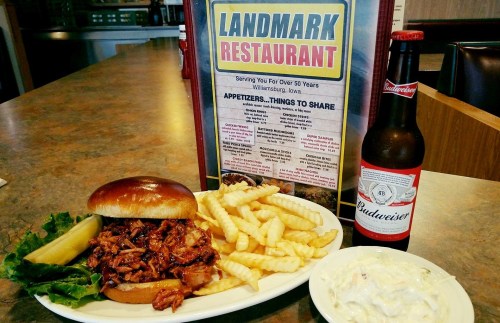 A plate featuring a pulled pork sandwich, fries, coleslaw, a pickle, and a Budweiser, with a menu in the background.