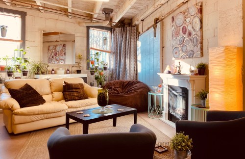 Cozy living room with a beige sofa, bean bag, fireplace, plants, and colorful artwork on the walls. Warm, inviting atmosphere.